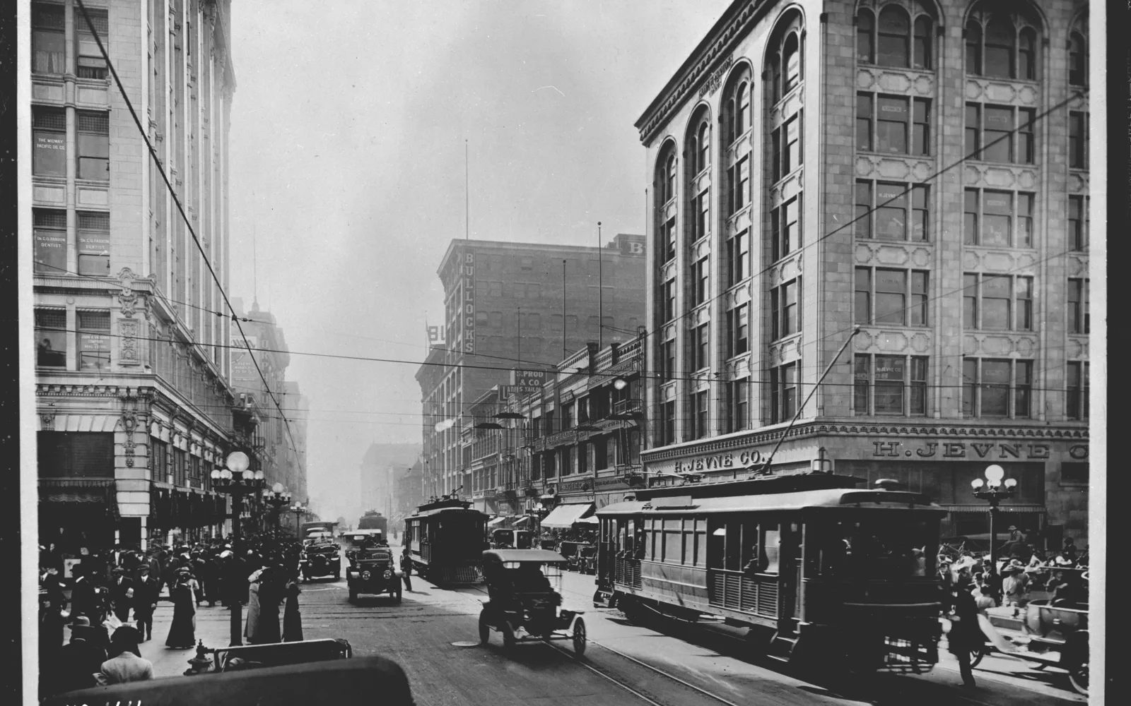 A historical street scene from Broadway in Los Angeles circa 1915, showing period storefronts, streetcars, and early twentieth-century urban architecture